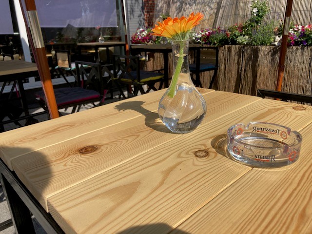 Zonnig terras van San's Place in Sint-Job-in-'t-Goor met houten tafels, verse gerbera's en kleurrijke bloembakken langs het rieten scherm